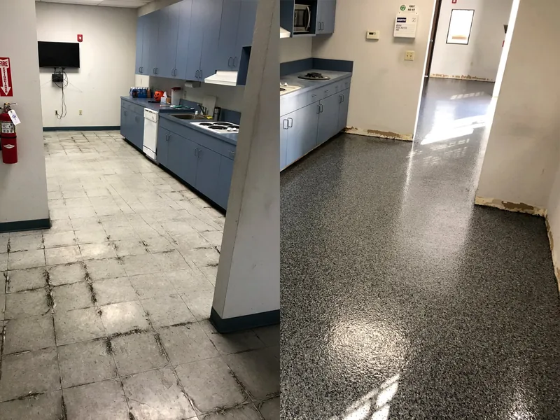 A break room with worn flooring beside its finished dark speckled floor and blue kitchenette.
